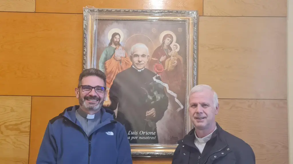 Padre Fernando (izquierda) y padre Mauricio (derecha) junto al cuadro de Don Orione. La llama en su mano representa las lágrimas de los pobres transformada en fuego y las espinas que brotan en una rosa, las dificultades.